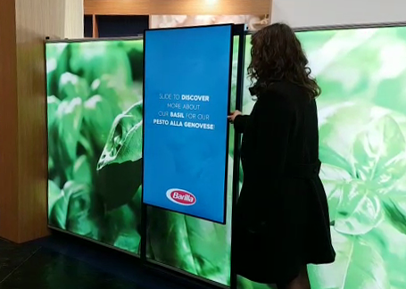 donna che utilizza la postazione multimediale dotata di un monitor scorrevole realizzata da ett presso lo stand di barilla all'evento cibus, sul monitor si vedono le foglie di basilico