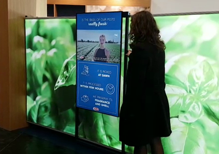 donna che utilizza la postazione multimediale dotata di un monitor scorrevole realizzata da ett presso lo stand di barilla all'evento cibus, sul monitor si vedono le foglie di basilico