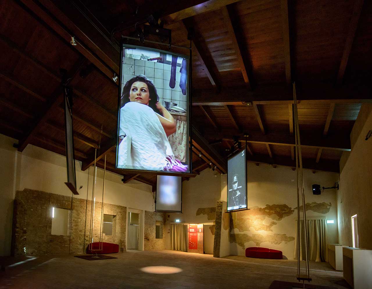 Una sala del Museo Fellini a Rimini con schermi, che riproducono celebri scene felliniane, scendono dal soffitto di travi a vista in legno, allestimento a cura di ett