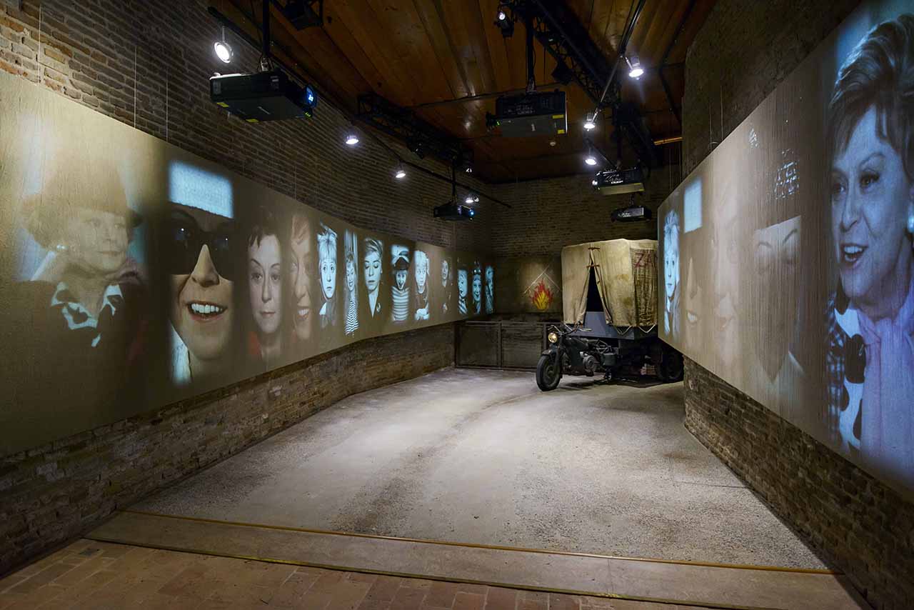 Una sala del Museo Fellini a Rimini, sulle pareti enormi schermi che riproducono attori in celebre scene di film felliniani, in fondo alla sala la riproduzione di un mezzo di trasporto (moto con rimorchio), allestimento a cura di ett