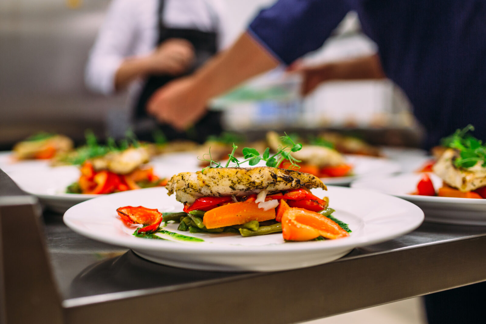 Piatto di pollo con verdure su un pass della cucina di un ristorante con cuochi sullo sfondo, utilizzata per la campagna bis contro gli sprechi alimentari, firmata da ett