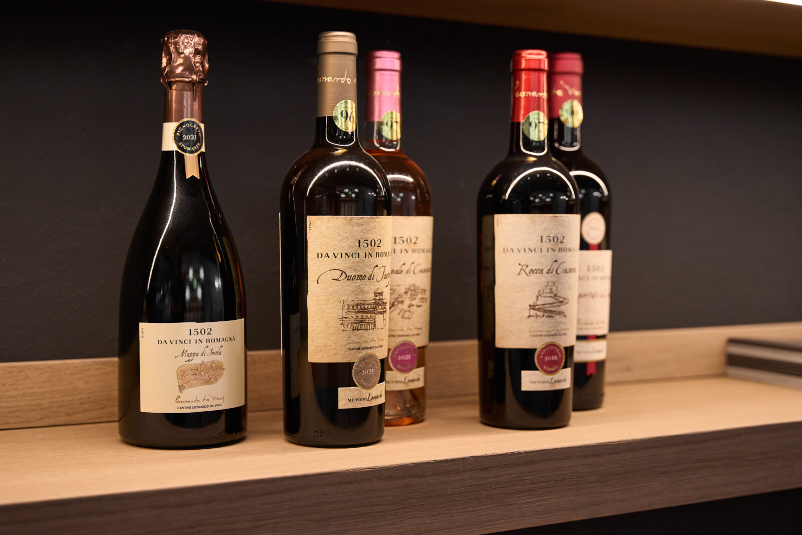Mensola in legno con sopra cinque bottiglie di vino nella galleria che ospita la mostra dedicata a leonardo da vinci e il suo rapporto con il vino, realizzata da ett