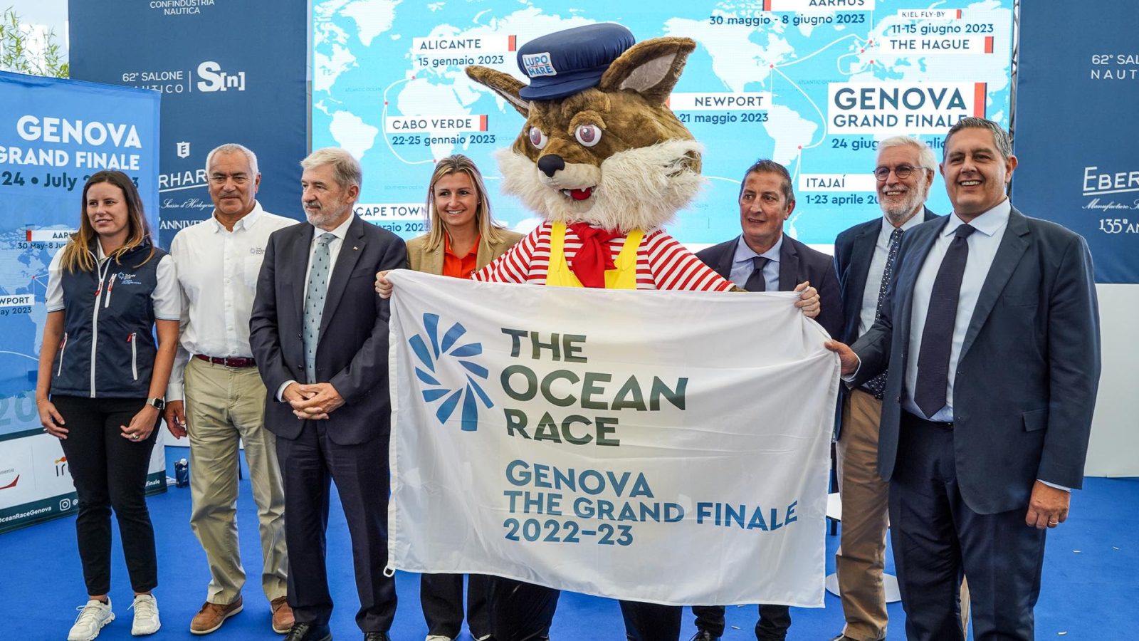 lupo di mare stringe la bandiera di the ocean race the grand finale di genova assieme a illustri personaggi genovesi