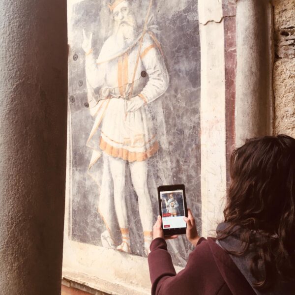 foto di una ragazza che utilizza l'applicazione del museo di palazzo besta osservando sul proprio cellulare la descrizione del grande affresco nel cortile interno davanti a lei