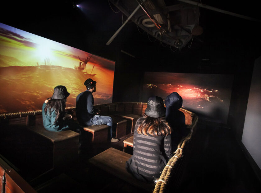 foto all'interno della sala della tempesta nel museo del mare a genova, tre visitatori su una barca riprodotta durante una tempesta