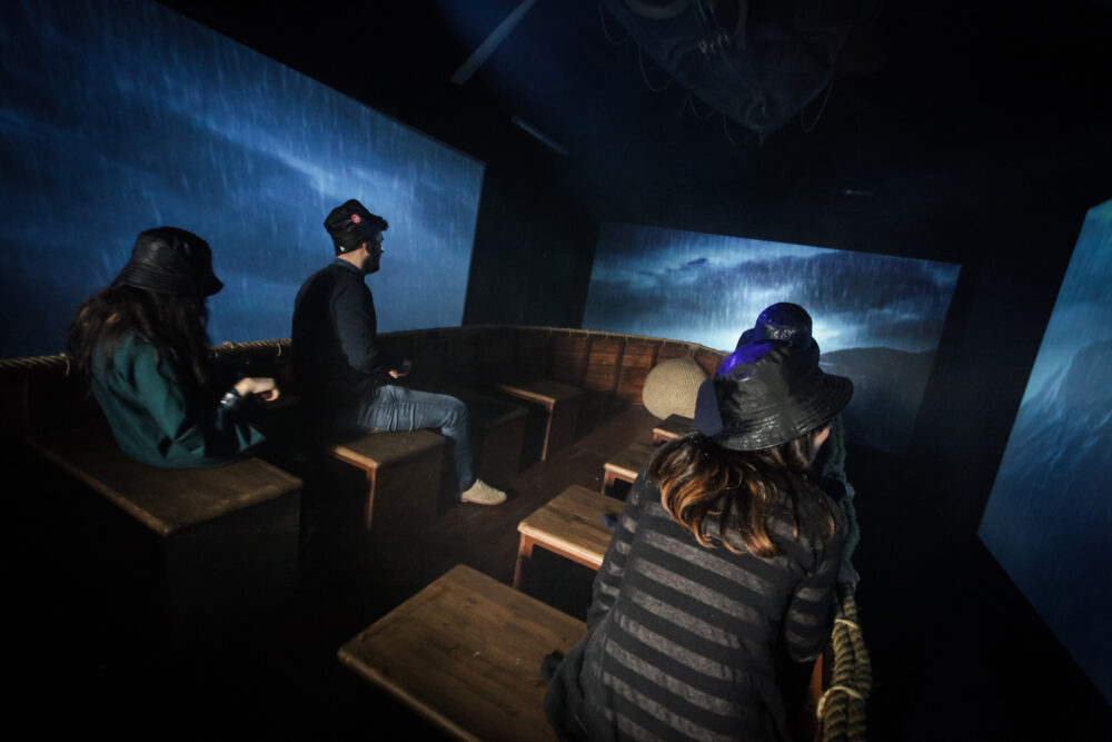 foto all'interno della sala della tempesta nel museo del mare a genova, tre visitatori su una barca riprodotta durante una tempesta, circondati dalla pioggia e monitor che simulano un mare agitato