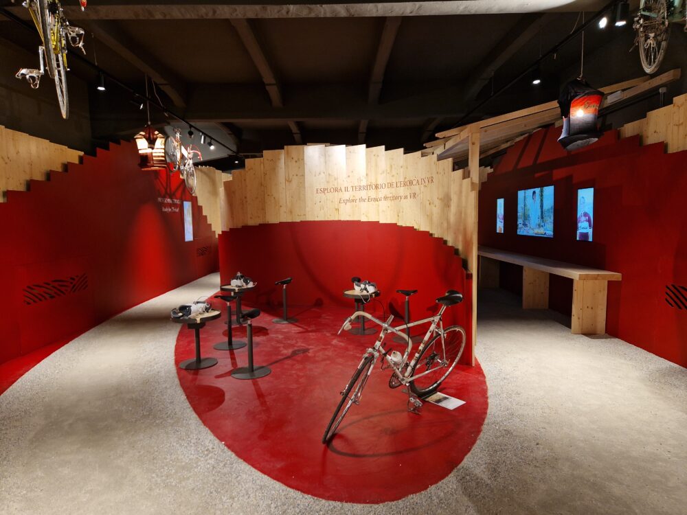 stanza di casa eroica, al centro della stanza una zona delimitata di rosso con una bici e alcune postazioni vr con visori