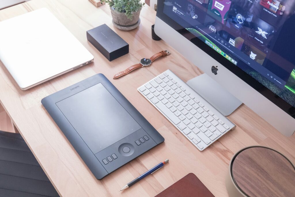 foto dall'alto di una scrivania in legno con un computer mac, un orologio da polso, un computer portatile chiuso, una penna e un tablet