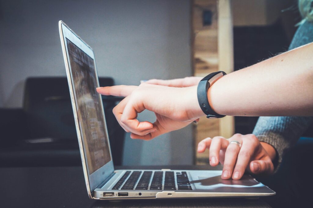 computer portatile su un tavolo nero fotografato di profilo aperto, mentre una mano clicca sul touchpad mentre la mano di una seconda persona indica lo schermo