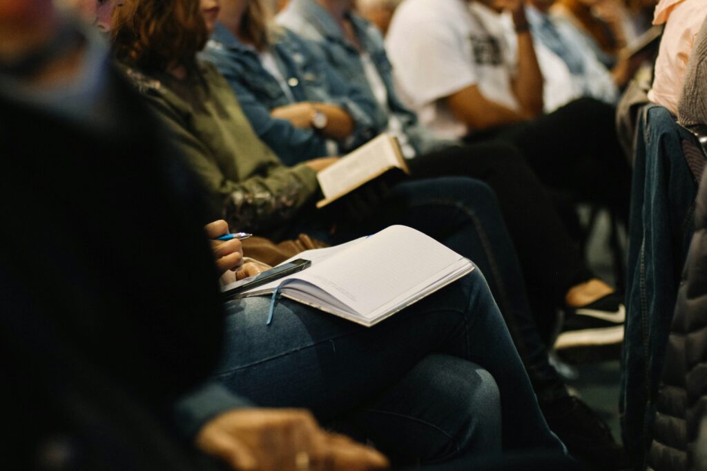 foto con inquadratura bassa sulle gambe di persone sedute durante una conferenza, alcuni con delle agende sulle gambe per prendere appunti