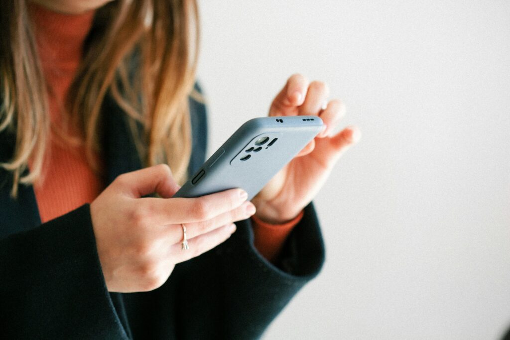 ragazza bionda con dolcevita arancione e cappotto nero con in mano un cellulare, mentre sta accedendo ai servizi digitali della PA