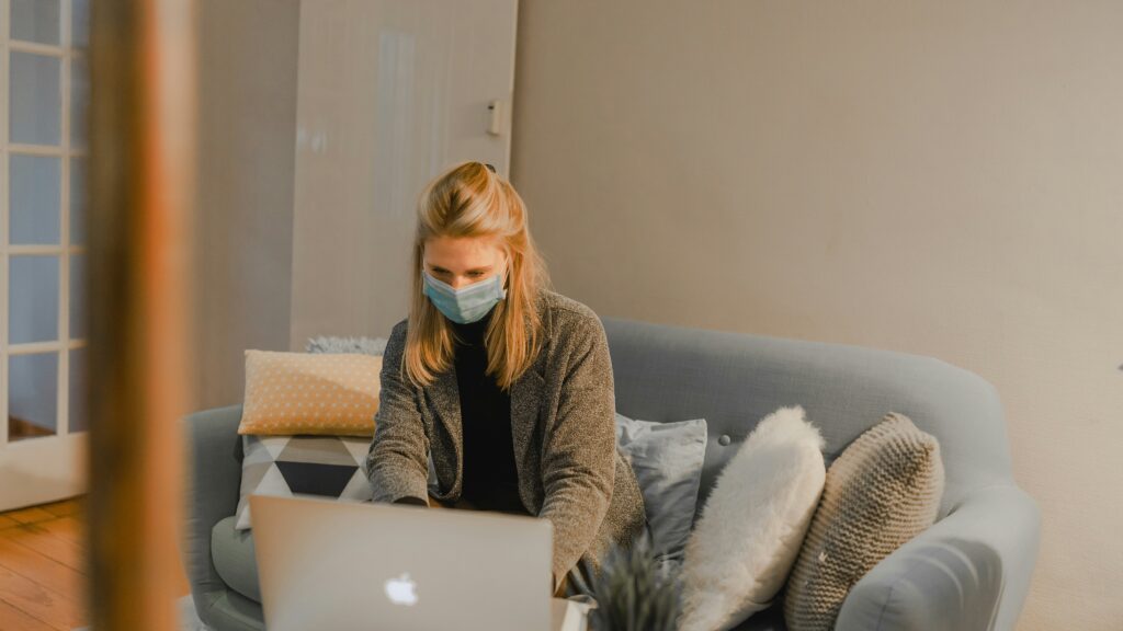 ragazza con la mascherina seduta sul divano di casa con davanti un computer portatile mentre fa smart working