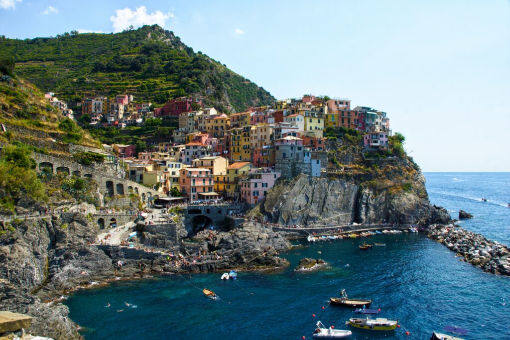 vista dal mare la costa di monterosso in liguria una delle cinque terre, le case colorate a picco sul mare costruite sulla roccia, alcune barche attraccate davanti al porticciolo e il sentiero pieno di persone