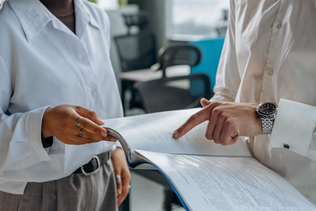 foto all'interno di una stanza d'ufficio di due busti: un signore, con al polso un orologio, sulla destra indica dei documenti che tiene all'interno di una cartellina, mentre una ragazza girata verso di lui regge i fogli, entrambi con la camicia