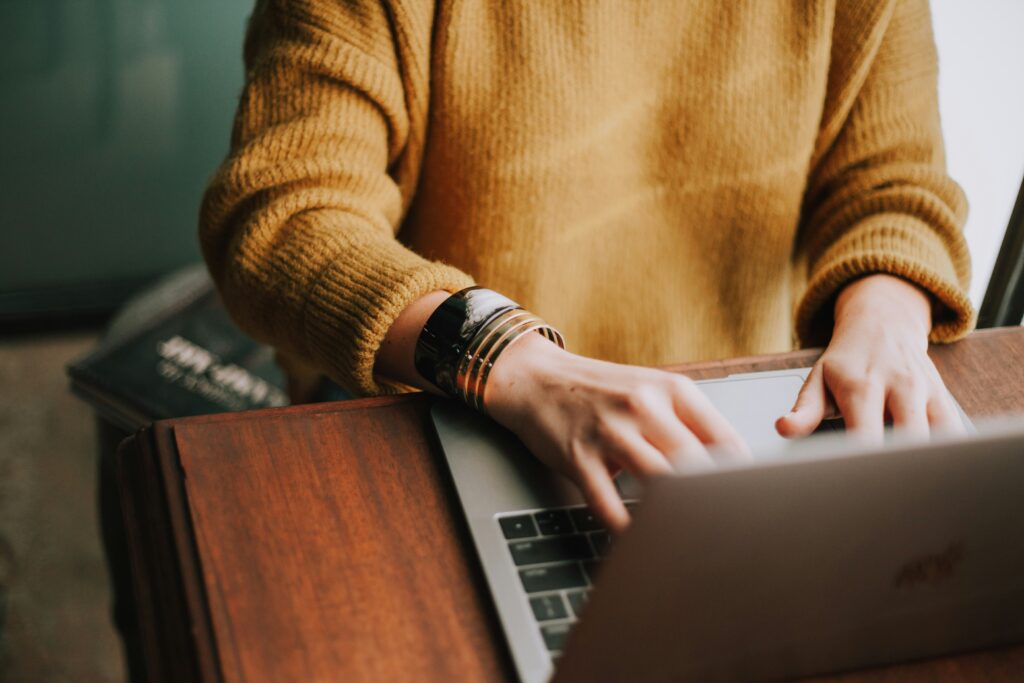 mezzobusto di una donna, con un maglione giallo ocra e due grandi bracciali, seduta davanti alla scrivania in legno che scrive sulla tastiera di un pc per prenotare online i CPI della campania