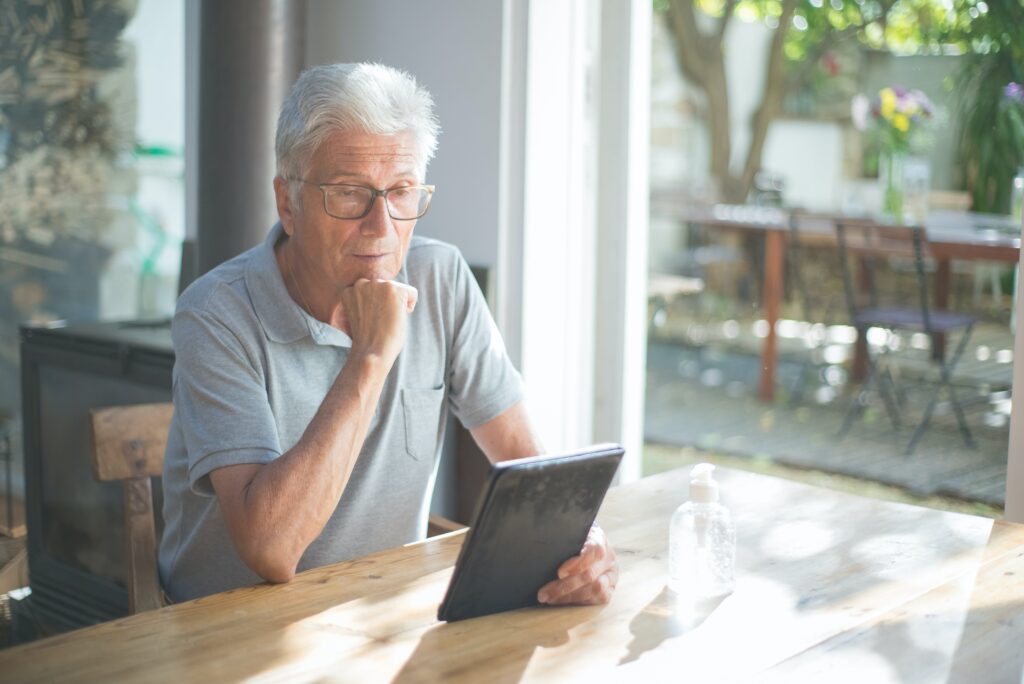 signore brizzolato con gli occhiali che indossa una polo grigia seduto ad un tavolo in legno accanto a una grande vetrata, che osserva il tablet che tiene in mano, sul tavolo davanti a lui il contenitore con l'igenizzante