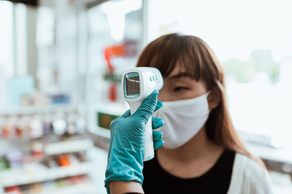 mano con guanto in lattice azzurro che misura la temperatura ad una ragazza con la frangetta che indossa la mascherina grazie a un termometro digitale, all'interno di una farmacia