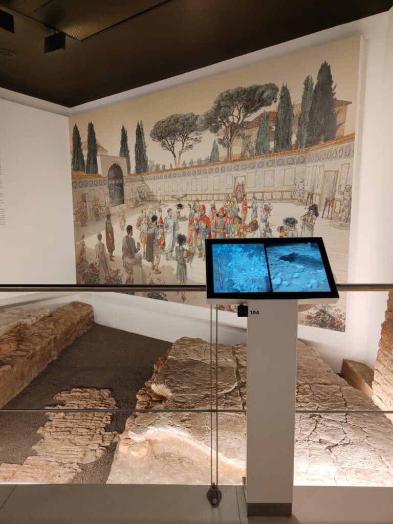 Sala con schermo su piedistallo presso il Museo del Ninfeo a Roma, allestimento museale di ett, sullo sfondo delle rovine, dei reperti antichi e sulla parete un'illustrazione di epoca romana