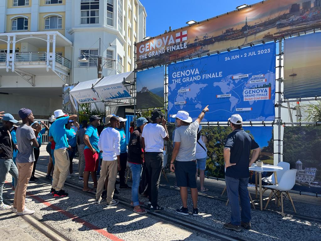 Un gruppo di persone osserva un cartellone che segue il percorso della gara intorno al mondo, con la proiezione delle tappe della regata sino all’arrivo a Genova per the ocean race Grand Finale