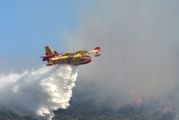 foto di un aereo giallo e rosso della securitè civile mentre è intento a gettare acqua per spegnere un incendio sullo sfondo della foto