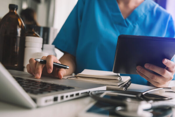 foto di un medico alla scrivania inquadrato, senza volto, intento a compilare il diario clinico con davanti un computer portatile dei block notes, in mano una penna e nell'altra un tablet, sullo sfondo dei flaconi di medicinali