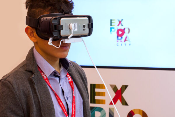 ragazzo con addosso il visore nello stand del progetto explora city, sullo sfondo un monitor con un video di explora city da far partire