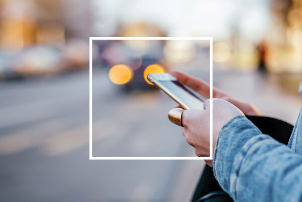 foto delle mani di una ragazza, sul ciglio della strada, con addosso una giacca di jeans e un grande anello color oro che tiene in mano un iphone mentre messaggia, sullo sfondo di un paesaggio urbano sfocato alle luci del tramonto