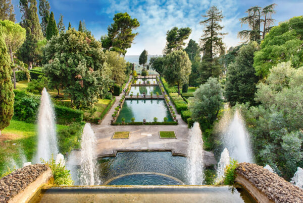 foto di un parco di una villa con al centro in primo piano una fontana e delle piscine d'acqua al centro del viale principale, intorno diverse varietà di alberi verdi