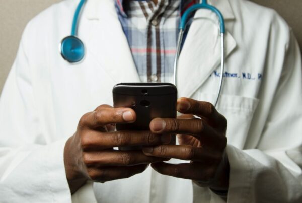 foto del busto di un medico che tiene al centro e in primo piano tra le mani un cellulare, con al collo un fonendoscopio