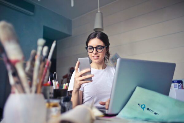 ragazza con occhiali alla scrivania davanti al computer portatile mentre guarda il cellulare, in primo piano sulla sinistra pennelli e colori per dipingere fuori fuoco