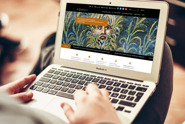 mockup su un macbook air del sito pompeii per il progetto tsp pianificazione percorsi turistici, che ha come schermata principale un mosaico di un volto sulle sfumature del verde, il pc è sulle gambe incrociate di un uomo seduto, in primo piano le sue mani che toccano la tastiera