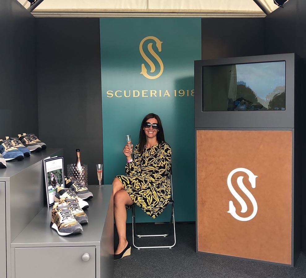 donna con occhiali da sole felice con bicchiere di spumante nello Showroom della scuderia 1918, sulla sinistra esposizione di scarpe, sulla destra monitor interattivo spento con sotto logo aziendale, nuova shopping experience realizzata da ett