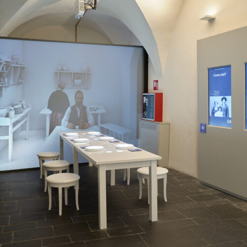 sala al museo dell'emigrazione italiana (MEI) con al centro un tavolo apparecchiato e sulla parete in fondo la proiezione immersiva di un uomo a capotavola che cena