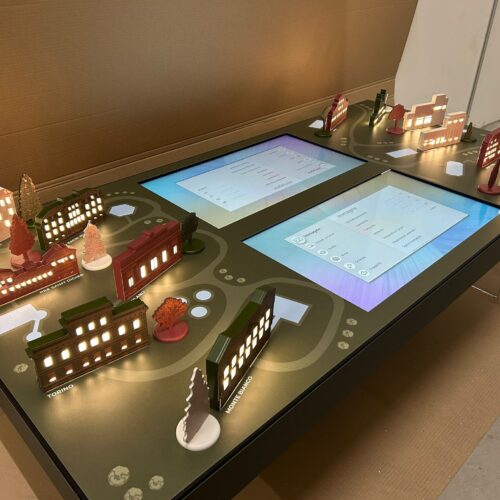 12 miniature 3d rappresentative dei diversi centri QC Terme disposte su un tavolo touch interattivo al centro del quale si trovano due monitor