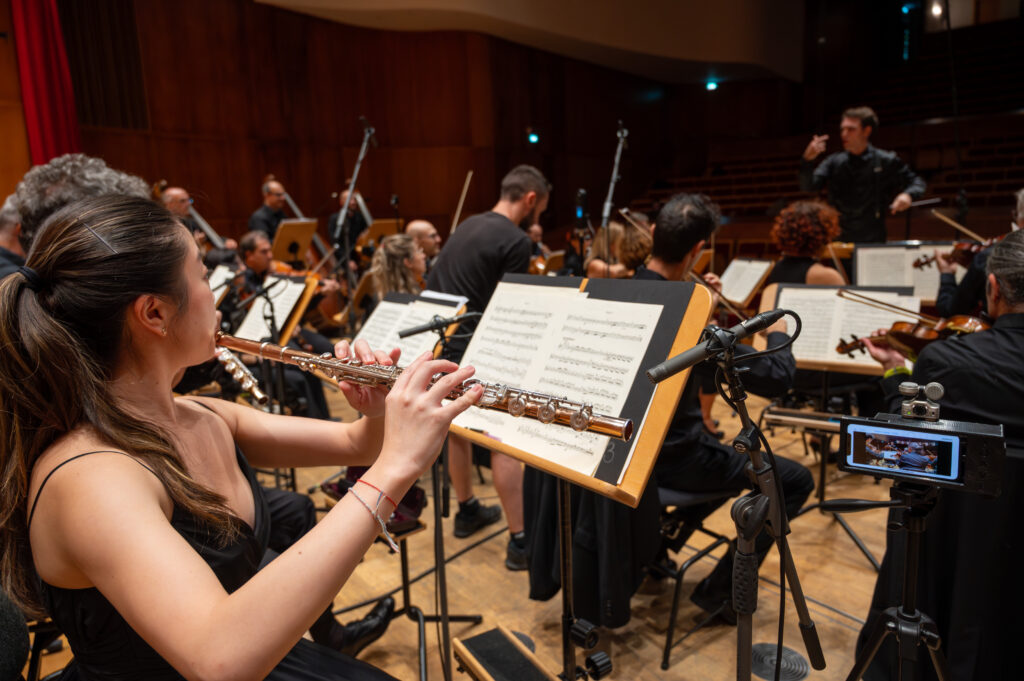 foto del backstage delle riprese a 360 durante un concerto sinfonico, nell'immagine abbiamo il punto di vista dei musicista dell'orchestra, fa parte del progetto SOAVE realizzato da ett. in primo piano di schiena una donna che suona il flauto traverso all'interno dell'orchestra